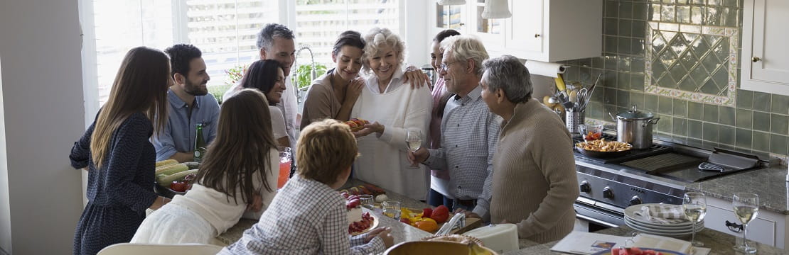 A group of 11 people gathers around a kitchen counter topped with serving plates full of food.